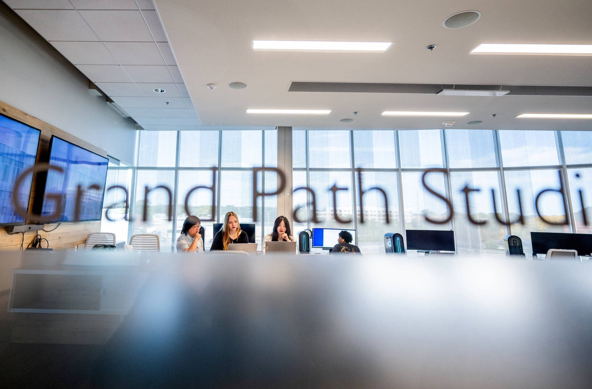three people working closely on computers in the background, Grand Path Studio door sign appears as if it's written above them on windows